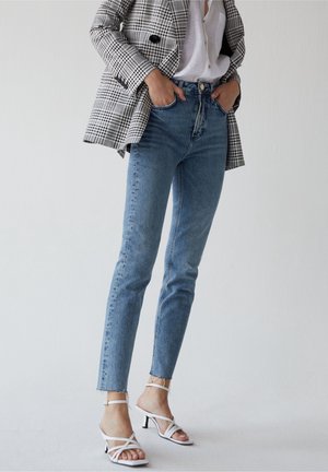 Osoba ubrana w niebieskie dżinsy z wysokim stanem, białe sandały na obcasie z paskami, białą koszulę na guziki oraz czarno-białą kratowaną marynarkę.