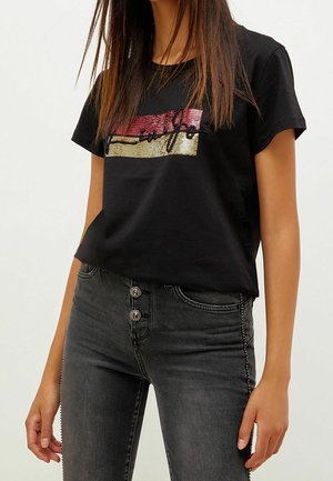 Kvinde iført en sort t-shirt med korte ærmer med røde og gyldne palietstriber og tekst, kombineret med højtaljede, mørkegrå jeans.