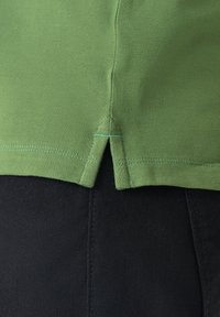 Zöld pamut póló, texturált felülettel, rövid oldalsó hasítékokkal az alján, és kontrasztos türkiz varrással. Fekete anyagra húzva viselve.