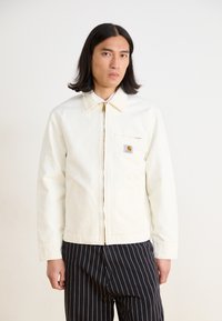 Witte canvasjas met kraag, voorrits en linkervak op de borst. Voorzien van een klein, gemerkt logopatch en een ontspannen pasvorm. Gestreepte broek.
