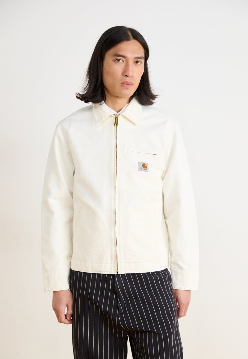 Witte canvasjas met kraag, voorrits en linkervak op de borst. Voorzien van een klein, gemerkt logopatch en een ontspannen pasvorm. Gestreepte broek.