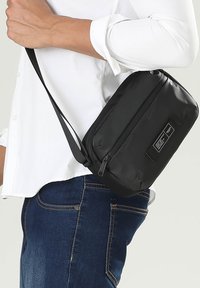 Borsa a tracolla nera realizzata in materiale texturizzato, con chiusura a zip, tracolla regolabile e una piccola etichetta sul fronte.