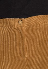 Gros plan sur la ceinture d'un pantalon en velours côtelé couleur fauve avec un seul bouton marron et un haut côtelé noir au-dessus.