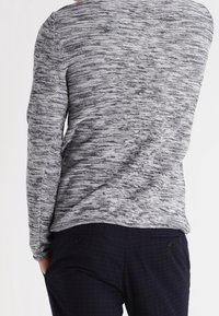 Maglione grigio con un design a maglia testurizzato, maniche lunghe e un ampio scollo, abbinato a pantaloni scuri su misura con sottili motivi a quadri.