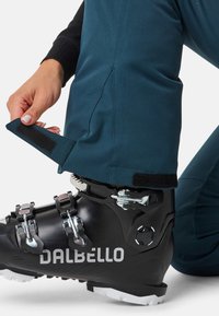 Schwarzer Skischuh mit silberner Hardware, ausgestattet mit verstellbaren Riemen. Teal-farbene Stoffhose mit Klettverschluss am Saum, die den Schuh sichtbar macht.