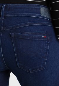 Lähikuva tummansinisistä Tommy Jeans -farkuista, jossa on takatasku, jossa on pieni punavirallinen ja sinivalkoinen lippulogon ja nahkabrändin merkki vyötäröllä.