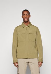 Veste vert olive clair avec fermeture éclair à l'avant, deux poches poitrine, un col, et une finition lisse et texturée, portée sur un pantalon beige.
