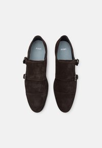 Chaussures en suede marron foncé avec deux sangles à boucle, dotées d'un design cousu et d'une texture lisse. L'intérieur est muni d'une semelle bleue portant l'inscription "JOOP !"