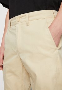 Beige maßgeschneiderte Hose mit glatter Textur, Gürtelschlaufen, Knopfverschluss und Seitentaschen, teilweise dargestellt mit einer Hand in der Tasche.