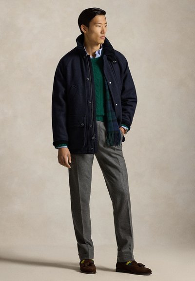 Marinblå jacka med hög krage, grå byxor, grön kabelstickad tröja och bruna loafers. Innehåller fickor och en mönstrad scarf.