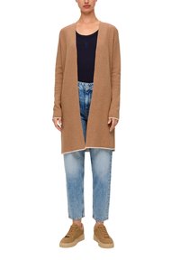 Long cardigan ouvert en beige, associé à un haut bleu marine et un jean clair déchiré. La chaussure choisie est des baskets beige avec des détails texturés.