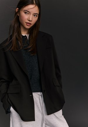 Mlada ženska z dolgimi rjavimi lasmi, oblečena v črni blazer, temen pulover in svetlo sive trenirke, na temni podlagi.