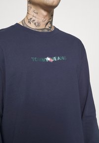 Mörkblå långärmad T-shirt gjord av mjukt tyg, med en grön broderad "TOMMY JEANS"-logotyp och en liten flaggdetalj på bröstet.