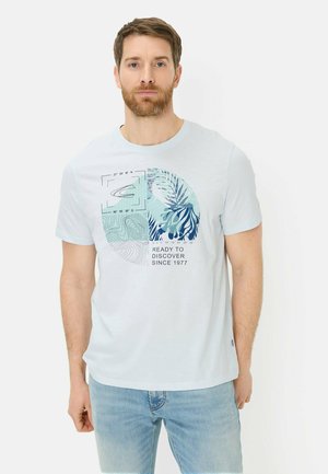 Mężczyzna z krótkimi brązowymi włosami i brodą, ubrany w jasnoniebieski t-shirt z nadrukiem mapy, liści i napisem "Gotowy do odkrywania od 1977."
