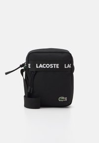 Black fabric crossbody bag featuring a white LACOSTE logo strap, top zipper closure, and a green embroidered crocodile logo on the front.