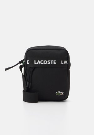Borsetta a tracolla nera in tessuto con una tracolla bianca con logo LACOSTE, chiusura con zip superiore e logo coccodrillo ricamato verde sul davanti.