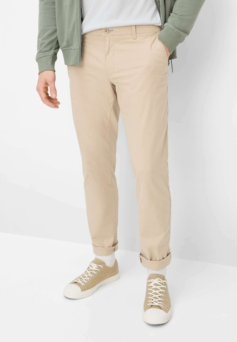 Beige katoenen chino's met een rechte pasvorm, voorzien van een opgerolde zoom, voorzakken en een subtiele knoopsluiting. Gecombineerd met lichtgekleurde sneakers.