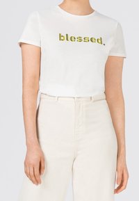 Vit bomulls t-shirt med korta ärmar, med glittrig gul text som säger "blessed." I kombination med off-white högmidjade byxor.