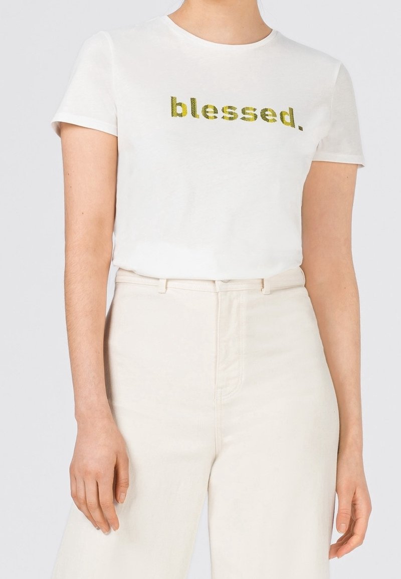 Vit bomulls t-shirt med korta ärmar, med glittrig gul text som säger "blessed." I kombination med off-white högmidjade byxor.