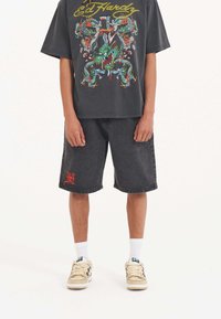 Black denim shorts with a red logo, paired with a black graphic t-shirt featuring colorful dragon designs. Worn with white socks and sneakers.