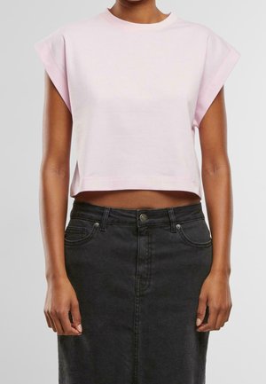 Nainen, jolla on vaaleanpunainen hihaton lyhyt toppi ja musta korkeavyötäröinen farkkuhame, seisoo yksiväristä taustaa vasten.