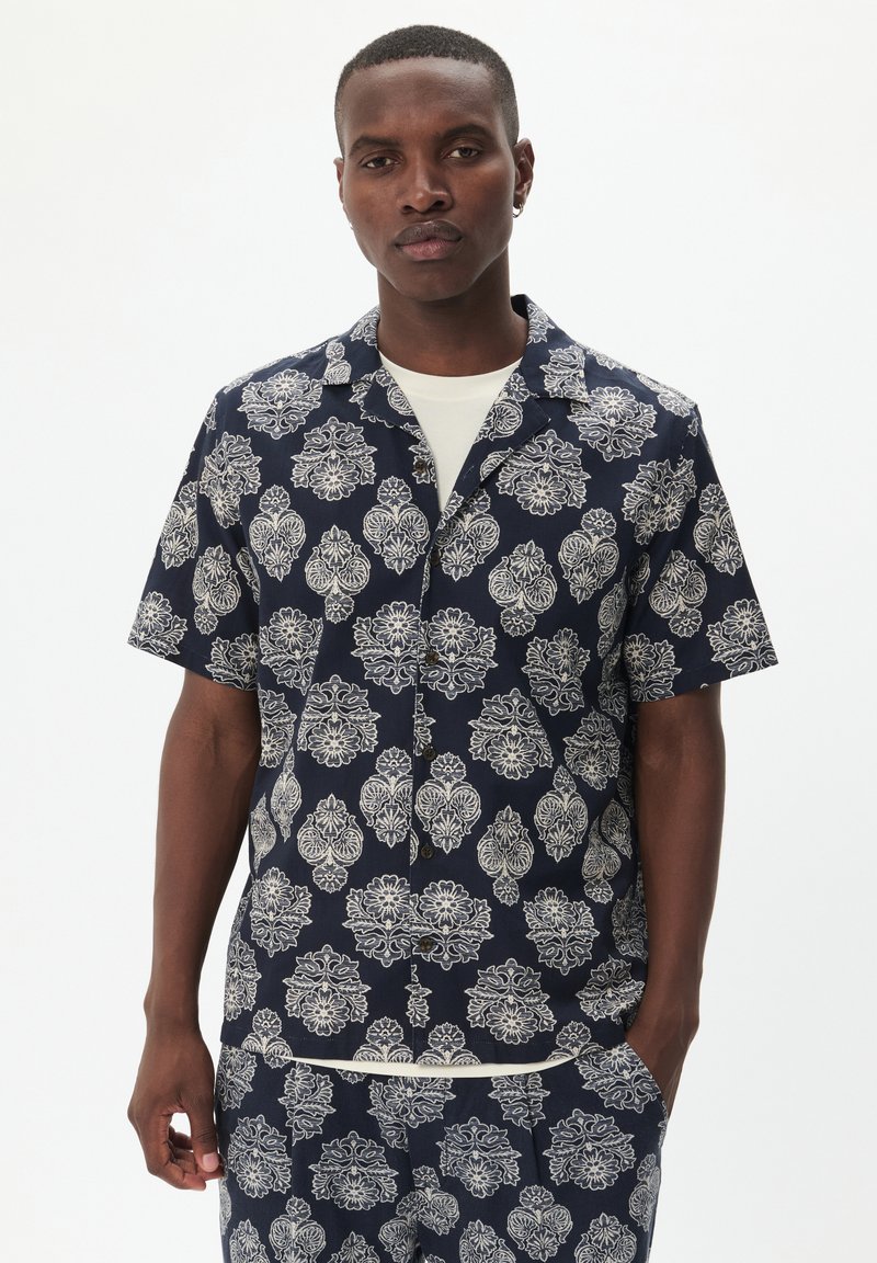 Camicia a maniche corte in tessuto blu navy con intricate fantasie floreali bianche, colletto a punte e chiusura frontale con bottoni.
