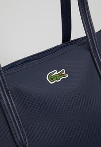 Sac à main bleu marine texturé avec doubles poignées et patch logo crocodile vert près de la fermeture éclair supérieure.
