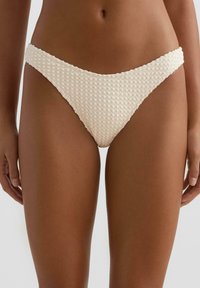 Ženska, ki nosi kremno barvne bikini spodnje hlačke z visokim izrezom in teksturiranim vzorcem vaflaste tkanine, prikazana od pasu do sredine stegna.