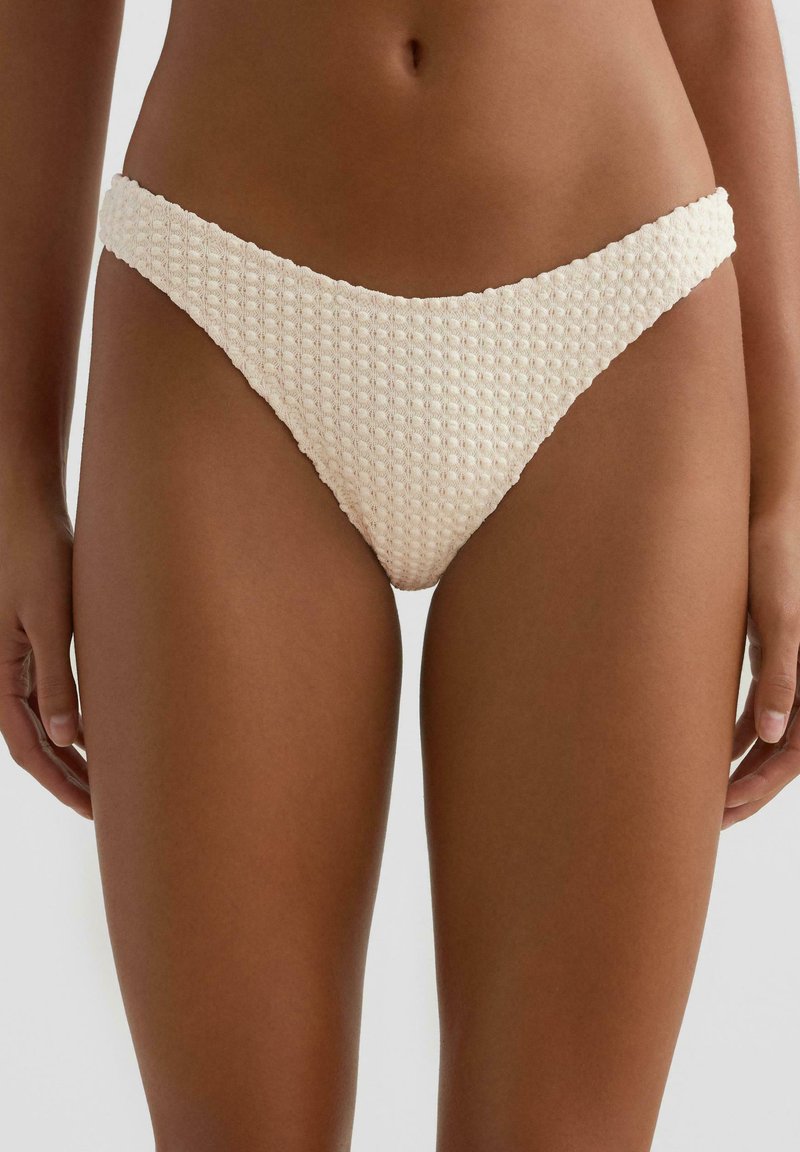 Ženska, ki nosi kremno barvne bikini spodnje hlačke z visokim izrezom in teksturiranim vzorcem vaflaste tkanine, prikazana od pasu do sredine stegna.