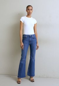 Maglietta bianca aderente abbinata a jeans a campana blu a vita alta, con un taglio pulito e design minimalista. Tacchi neri con strappi completano il look.