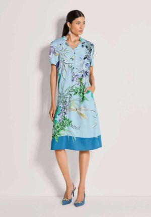 Hellblaues Blumenkleid mit kurzen Ärmeln, V-Ausschnitt und Taschen. Es verfügt über einen großen botanischen Druck und einen einfarbigen blauen Saum. Kombiniert mit blauen High Heels.