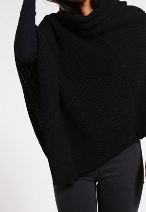 Kvinde iført en sort ribstrikket poncho med krave over en mørk langærmet top og mørke bukser mod en ensfarvet baggrund.