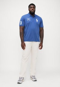 Tricou polo albastru din bumbac, cu guler, având un logo brodat alb și numărul 3 pe mânecă. Asortat cu pantaloni bej deschis și tenisi.