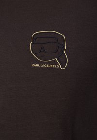 Schwarzes T-Shirt mit einer gestickten Grafik eines Charakters mit Sonnenbrille. Darunter steht in heller Schrift "KARL LAGERFELD".