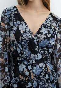 Sukienka w kwiaty z czarnym tłem, niebieskimi i beżowymi kwiatami, długimi rękawami, głębokim dekoltem w kształcie litery V oraz detalem w postaci zawiązanego pasa.