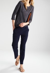 Marinblå, slim-fit byxor i kombination med en mörk topp med vita prickar. Personen bär bruna remsandaler och håller i en brun clutch.