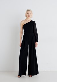 Moteris su trumpais šviesiais plaukais, dėvinti juodą viena petimi perrišamą jumpsuit'ą su viena ilga skaidraus audinio rankove ir plačiomis keln�ėmis, stovinti ant baltos fono.