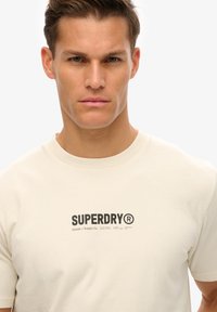 Camiseta de manga corta color crema hecha de algodón, con un logo y texto en negro de "SUPERDRY" en la parte frontal, cuello redondo y corte estándar.