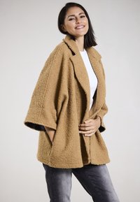 Tan, textured oversized outerwear with a shawl collar and half-length sleeves. Worn over a white top, paired with gray pants.