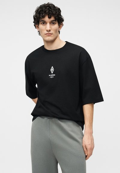 Νεαρός με σγουρά μαλλιά που φοράει μαύρο μεγάλου μεγέθους T-shirt με λευκό λογότυπο και γκρι παντελόνι, στέκεται μπροστά σε απλό φωτεινό φόντο.