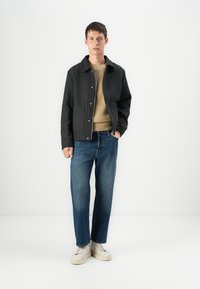 Veste à carreaux noirs, pull en tricot beige, jeans bleus et baskets blanches. La veste possède une fermeture à boutons et deux poches avant.