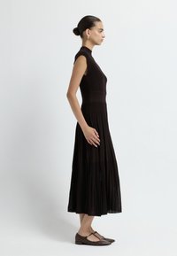 Černé, vroubkované, bezrukávové šaty s vysokým výstřihem a sklady na sukni. Modelka má na sobě hnědé, ploché, vzorované boty.