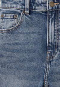 Gros plan sur la zone de la poche d’un jean en denim bleu avec des coutures orange et des rivets métalliques aux coins de la poche.