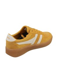 Zapatilla de ante amarilla con rayas y logo blancos. Presenta una suela de goma y un diseño de cordones. Parte superior texturizada y cuello acolchado para mayor comodidad.