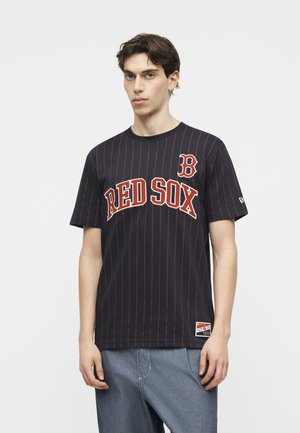 Mladý muž nosi čierne tričko Red Sox s tenkými pruhmi a svetlomodré rifle, stojaci pred jednoduchým bielym pozadím.