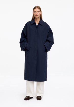 Femme portant un long manteau oversize bleu marine avec les mains dans les poches, pantalon blanc et chaussures marron foncé, debout devant un fond blanc uni.