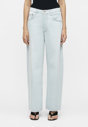 Γυναίκα με ανοιχτό μπλε τζιν παντελόνι με φαρδιά πόδια, μαύρο τοπ και μαύρα σανδάλια τύπου thong, στέκεται μπροστά σε απλό φωτεινό φόντο.