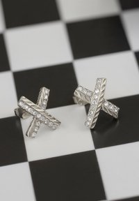 Silverörhängen med ett kors och tvärs mönster prytt med klara, glittrande stenar, mot en svart och vit rutmönstrad bakgrund.