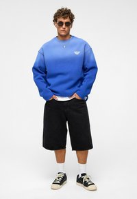 Blauwe gradient sweater met een wit logo, zwarte shorts en zwarte sneakers. Model draagt een zonnebril en gelaagde kettingen.