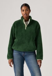 Γυναίκα που φοράει σκούρο πράσινο φλις μπλουζάκι με μισό φερμουάρ και ανοιχτόχρωμο τελείωμα, συνδυασμένο με μπλε τζιν, στέκεται μπροστά σε απλό φόντο.